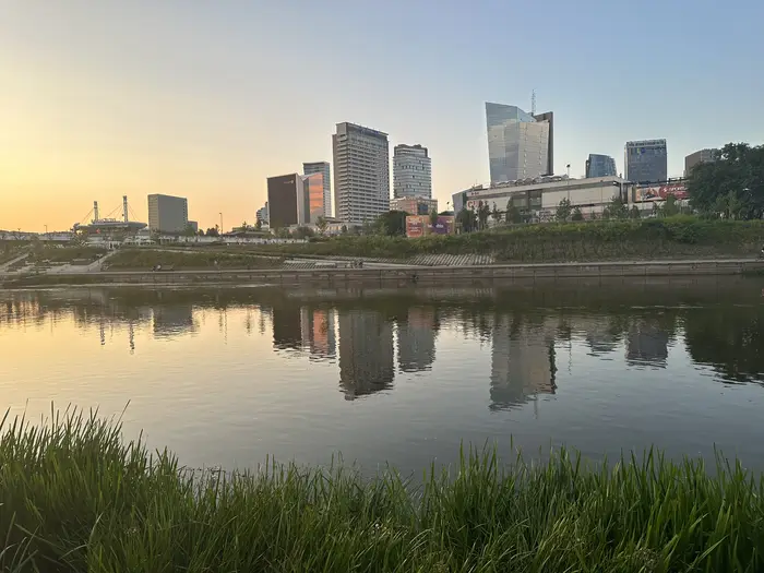 A view across the Neris River in Vilnius showing the modern skyline of high-rise buildings reflected in the water, framed by green grass in the foreground at sunset.