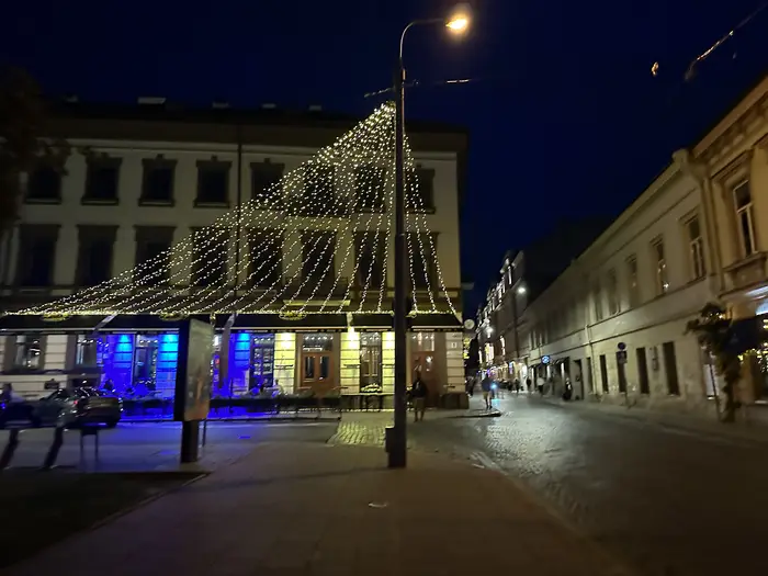 A night view of a cobblestone street in Vilnius Old Town, featuring a building decorated with draped string lights and a glowing street lamp.