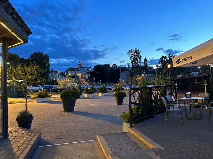 An outdoor cafe terrace with tables and chairs overlooks a paved plaza and distant historic church towers under a deep blue twilight sky in Vilnius.