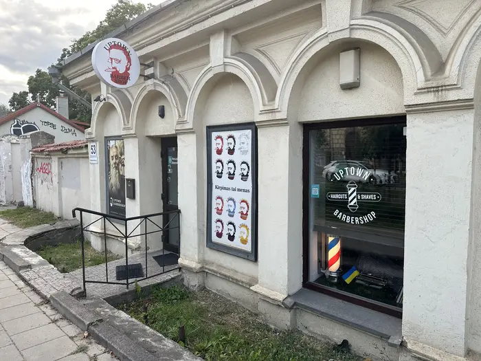 Exterior view of Uptown Barbershop in Vilnius, featuring arched windows, a circular logo sign, and posters on a light-colored stone building.