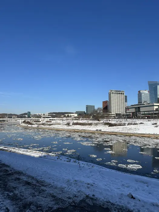 A winter view of the Neris River in Vilnius filled with floating circular ice floes, with snowy banks and modern skyscrapers under a clear blue sky.