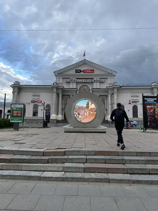 A large circular futuristic screen installation called 'The Portal' stands in the plaza in front of the Vilnius Railway Station, with a person walking towards it.