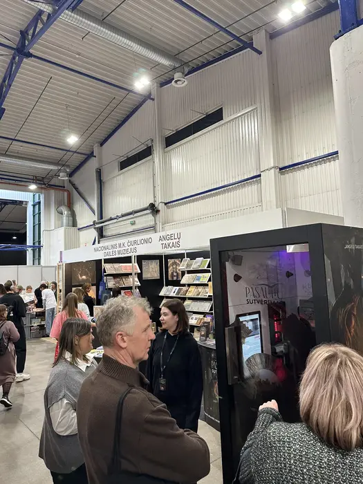 People gathered around an exhibition booth for the National M.K. Čiurlionis Art Museum inside a large exhibition hall.