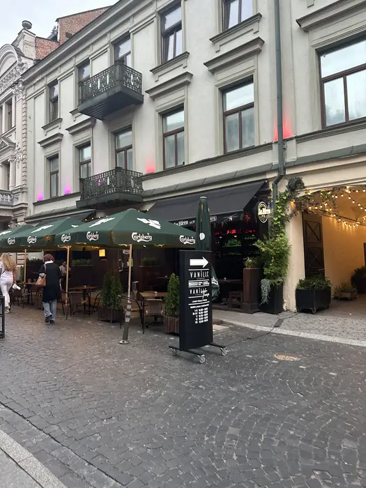 A cobblestone street in Vilnius featuring outdoor cafe seating under green Carlsberg umbrellas next to historic European buildings.