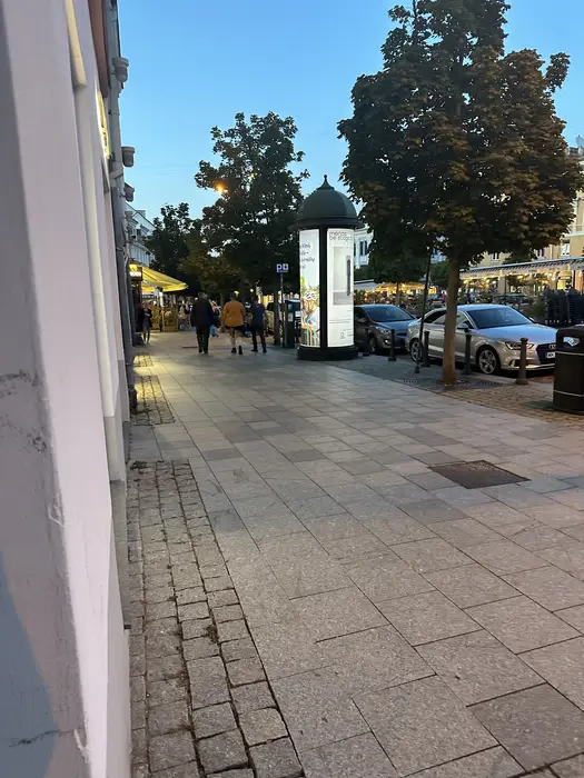 A wide stone-paved sidewalk in Vilnius at dusk, with people walking in the distance past trees and an advertising pillar.