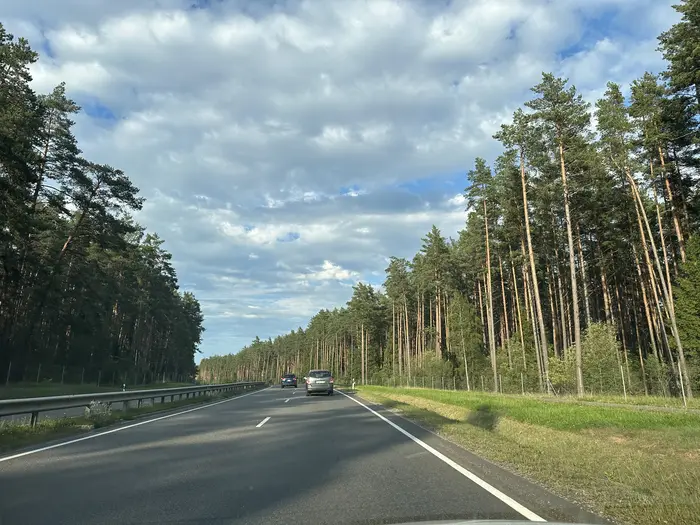 A two-lane asphalt highway stretching forward, flanked on both sides by tall, dense pine trees under a cloudy blue sky.