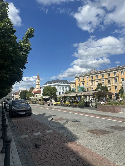 A cobblestone street in Vilnius Old Town with outdoor cafe seating, historic buildings, a church spire in the distance, and a parked car on the left under a blue sky with white clouds.
