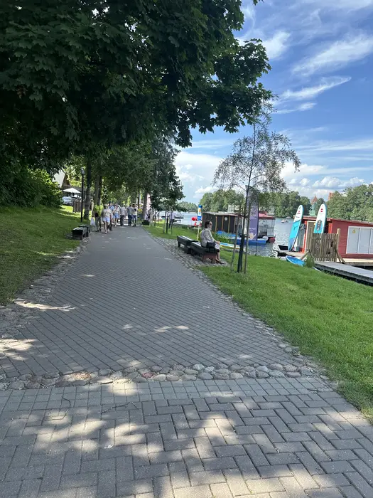 A paved pedestrian path lined with trees and benches runs alongside a lake with boats and paddleboards under a blue sky.