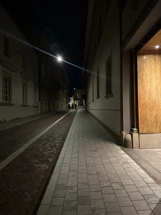 A narrow paved street in Vilnius Old Town at night, illuminated by a street lamp with a bright lens flare, showing historic buildings and distant figures walking.