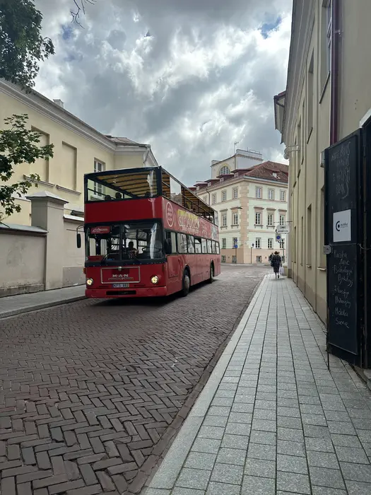 A red double-decker tour bus driving down a narrow cobblestone street lined with historic yellow buildings under a cloudy sky in Vilnius.