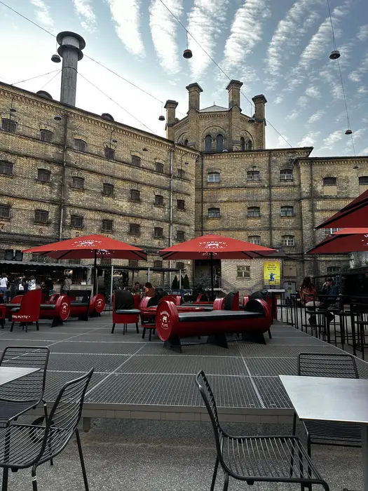 An outdoor patio with red umbrellas and barrel-shaped benches in the courtyard of the historic yellow-brick Lukiškės Prison in Vilnius.