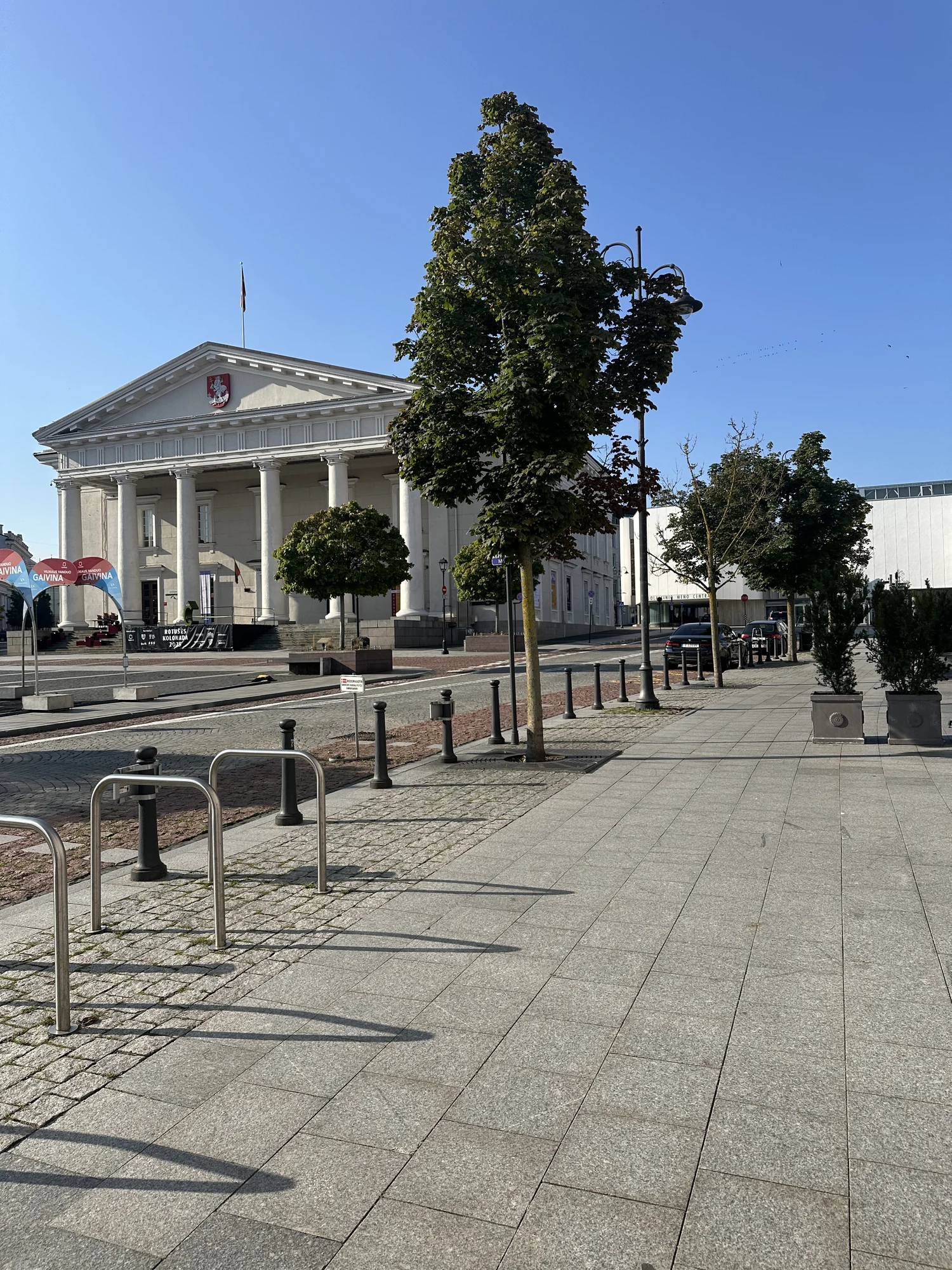 The neoclassical Vilnius Town Hall building seen from across the paved Town Hall Square on a sunny day with bike racks in the foreground.