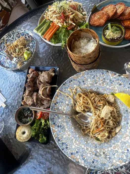 An overhead view of a glass table filled with various Thai dishes, including Pad Thai, papaya salad, grilled meat, and sticky rice in a bamboo basket.